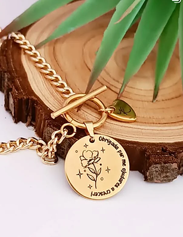 Pulseira dourada de elos com fecho de barra e uma medalha redonda gravada com uma flor e uma frase de agradecimento.