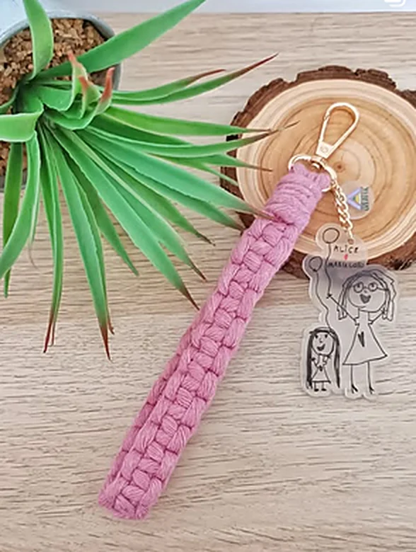 Porta-chaves com alça de macramé rosa e um pendente em acrílico transparente com o desenho de uma criança, recortado à volta.