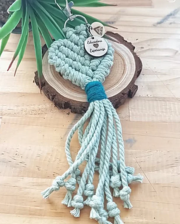 Porta-chaves em macramé verde-menta em forma de coração, com franja e medalha de madeira gravada, sobre uma fatia de tronco.